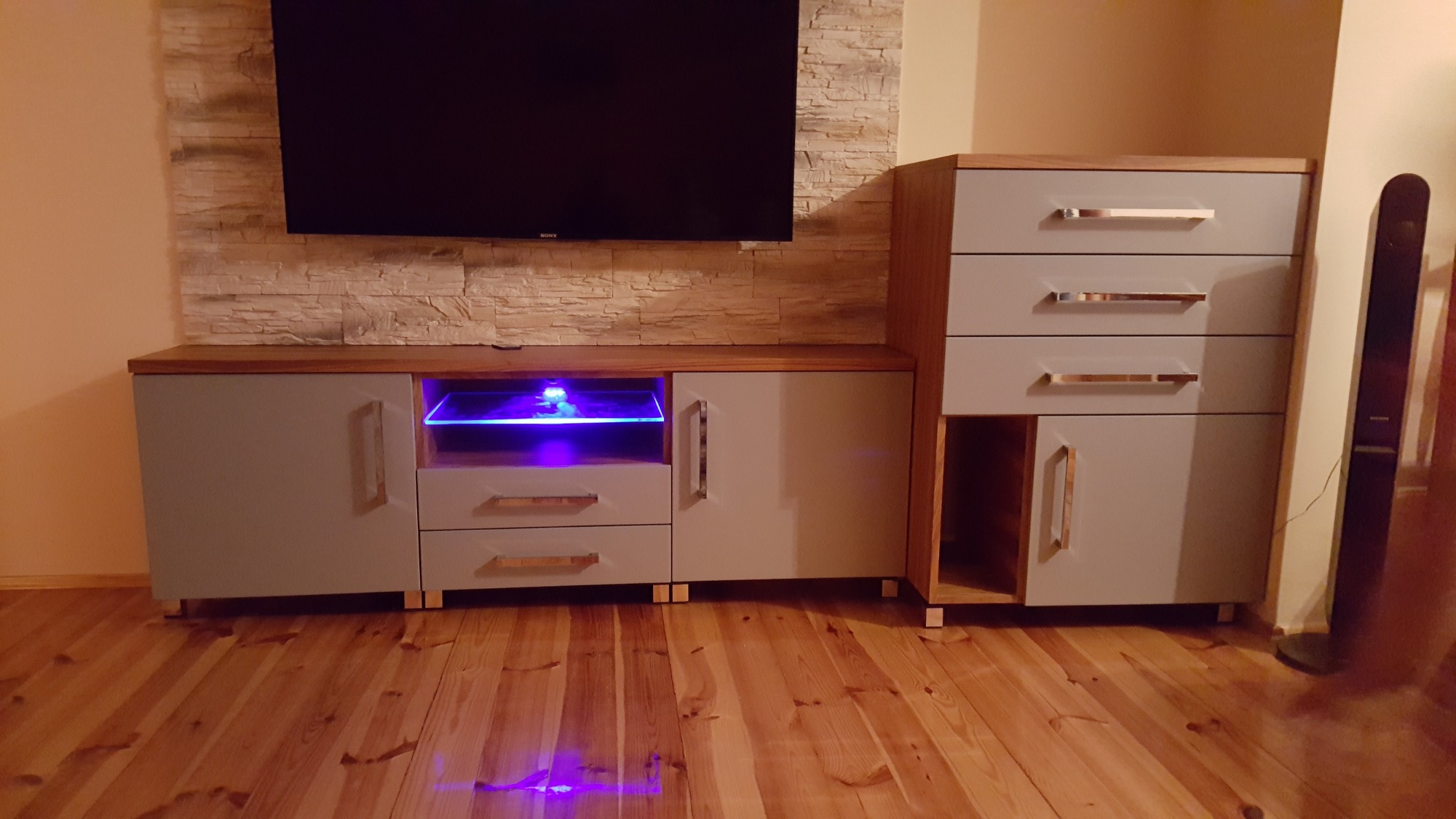 Zestaw mebli RTV z szarymi frontami, drewnianym blatem i podświetleniem LED, na drewnianej podłodze, z telewizorem na ścianie z imitacją kamienia.