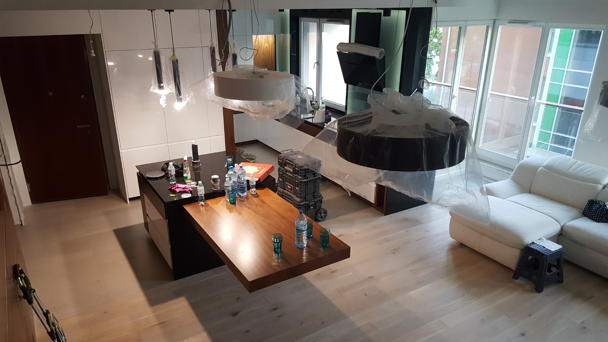 Wnętrze w trakcie remontu: wyspa kuchenna z czarnym blatem i drewnianym przedłużeniem, narzędzia i butelki na blacie, wiszące lampy zabezpieczone folią, biała sofa w tle, podłoga z jasnego drewna.