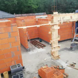 Wznoszenie ścian z czerwonej cegły na budowie domu, widoczne drewniane szalunki, drabina i pracownik w górze, na pierwszym planie mężczyzna z rurami PCV.