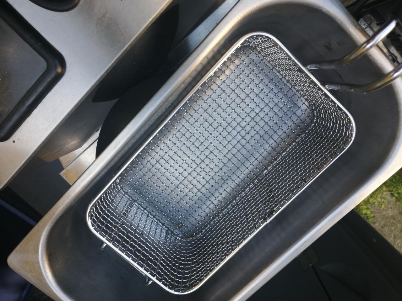 Kosz frytkownicy ze stali nierdzewnej wewnątrz zbiornika, widoczne pozostałości oleju, fragment blatu kuchennego w tle.