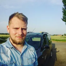 Młody mężczyzna w dżinsowej koszuli robi selfie na tle czarnego samochodu z napisem 'TAXI' na dachu, widoczne pole i budynki w tle.