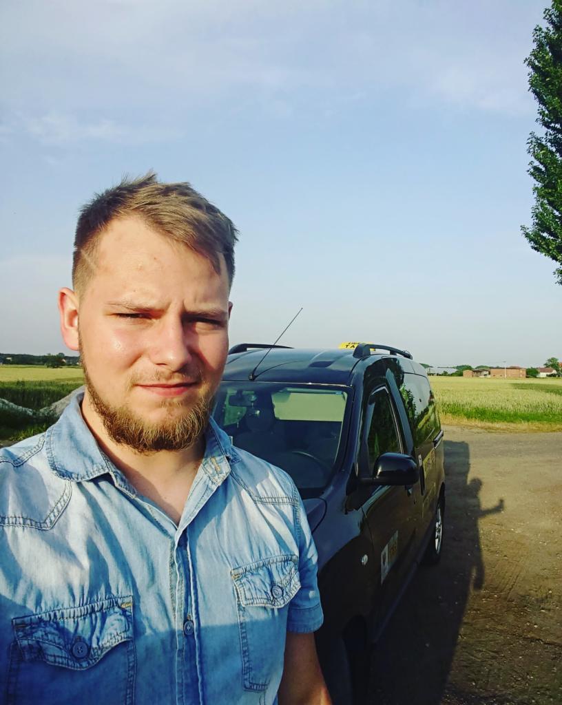 Młody mężczyzna w dżinsowej koszuli robi selfie na tle czarnego samochodu z napisem 'TAXI' na dachu, widoczne pole i budynki w tle.