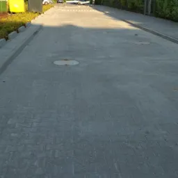 Ulica z szarej kostki brukowej w słoneczny dzień, widoczne studzienki kanalizacyjne, budynki w tle i przejście dla pieszych.