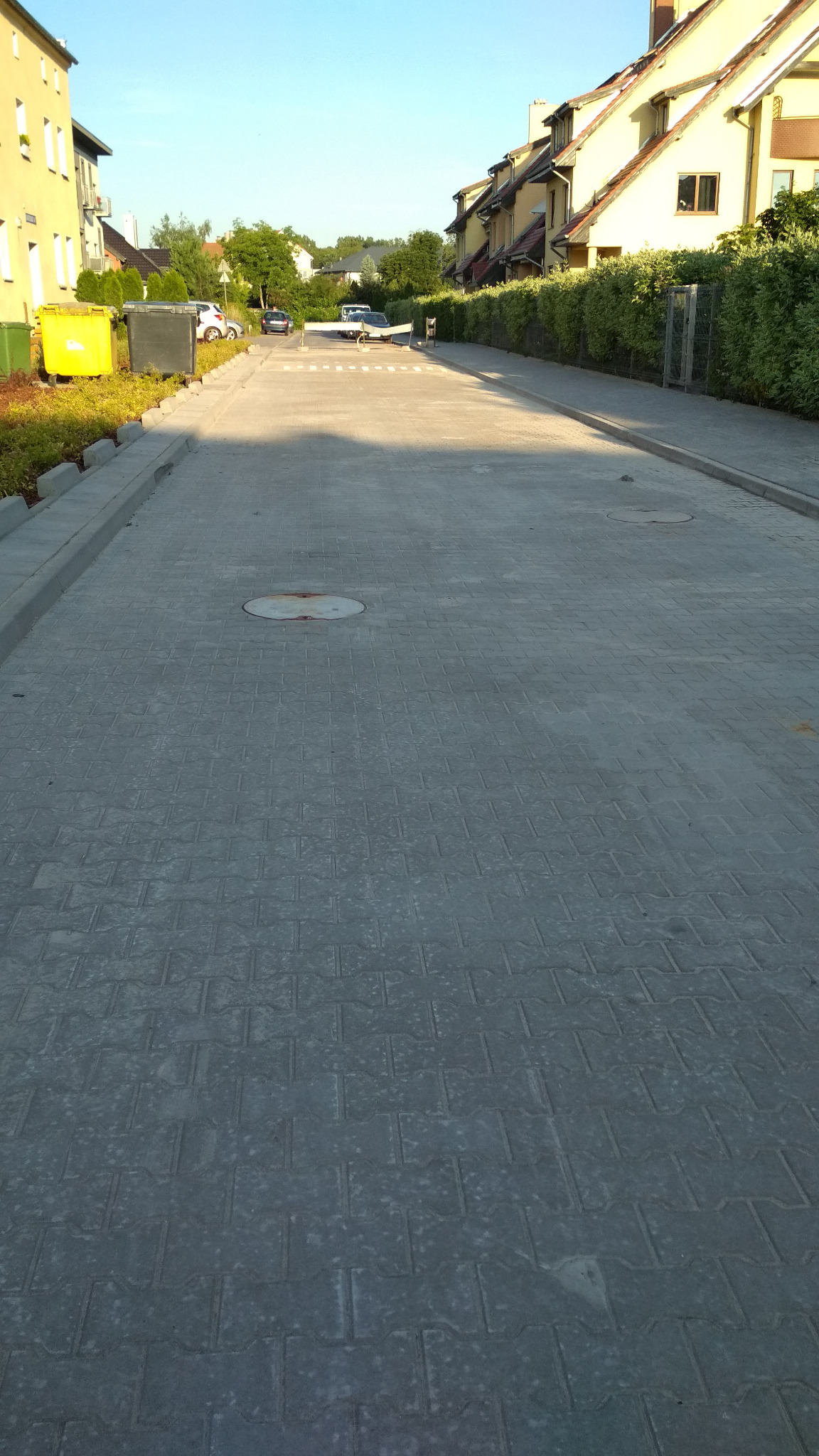 Ulica z szarej kostki brukowej w słoneczny dzień, widoczne studzienki kanalizacyjne, budynki w tle i przejście dla pieszych.