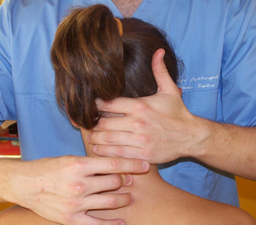 Fizjoterapeuta w niebieskim uniformie wykonuje masaż szyi pacjentki, widok z tyłu, skupienie na dłoniach i szyi, w tle fragment żółtej ściany.