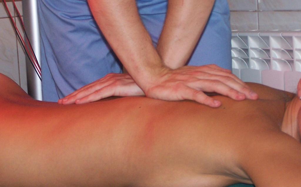 Masaż pleców pacjenta wykonywany przez terapeutę w niebieskim uniformie, widoczne dłonie terapeuty na plecach pacjenta, w tle białe kafelki i czerwone kable.