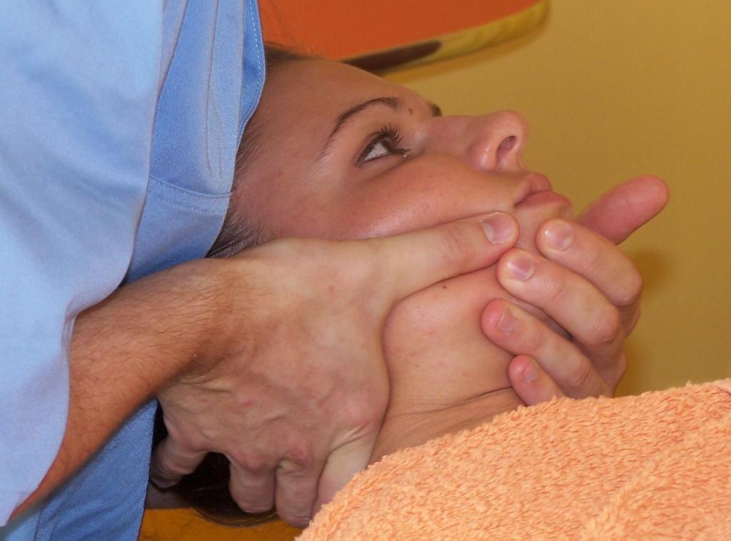 Kobieta leży na pomarańczowym ręczniku, podczas gdy terapeuta masuje jej okolice żuchwy i szyi