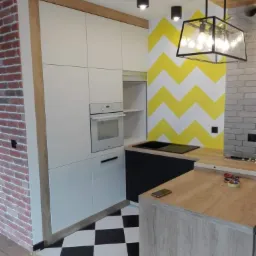 Nowoczesna kuchnia z białymi szafkami, czarnym blatem, podłogą w szachownicę i ścianą w żółto-białe zygzaki, oświetlona wiszącą lampą w stylu industrialnym.