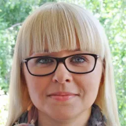 Portret kobiety z blond włosami i okularami w Poznaniu, uśmiechniętej i patrzącej w obiektyw, na tle zieleni.