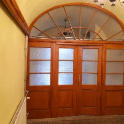 Drewniane drzwi wejściowe z półokrągłym przeszkleniem nad drzwiami, widoczne na tle żółtej ściany i grzejnika.