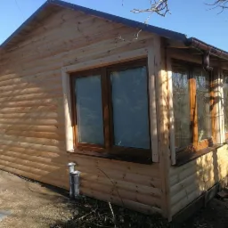 Nowo wybudowany, drewniany domek letniskowy z brązowymi ramami okiennymi i rynnami, widoczny w słoneczny dzień na tle drzew.