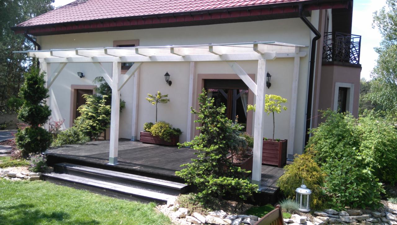 Biała pergola z drewnianym tarasem i stopniami przed domem, otoczona zielenią i kamieniami ozdobnymi, z widocznymi lampami na ścianie budynku.