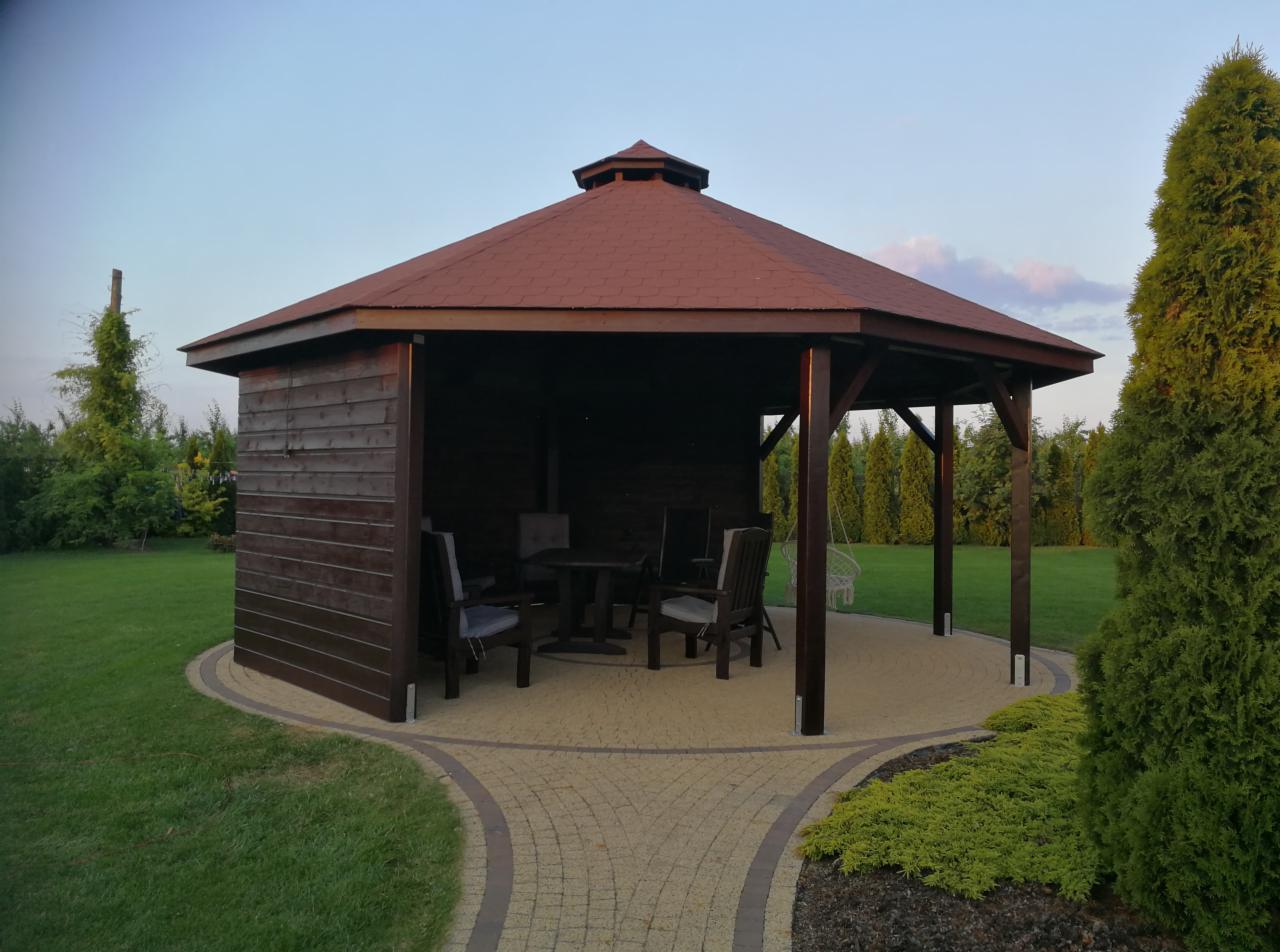 Ośmiokątna, ciemnobrązowa konstrukcja z drewna z czerwonym dachem, wyposażona w stół i krzesła, otoczona kostką brukową w kolorze piaskowym oraz zielonym trawnikiem i iglakami.