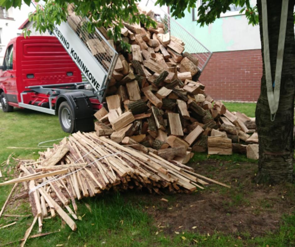 Ciężarówka z podniesioną skrzynią wyładowuje stos pociętego drewna na trawnik, obok leży związana wiązka cienkich gałęzi.