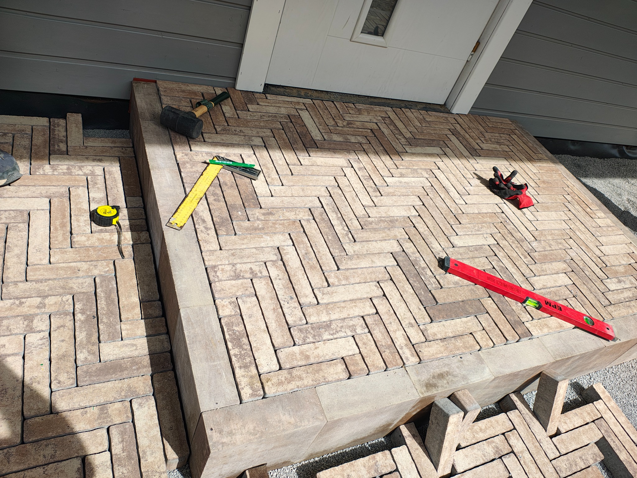 Ułożona kostka brukowa w jodełkę przed wejściem do domu, widoczne narzędzia: poziomica, młotek gumowy, miarka, kątownik i rękawice robocze.