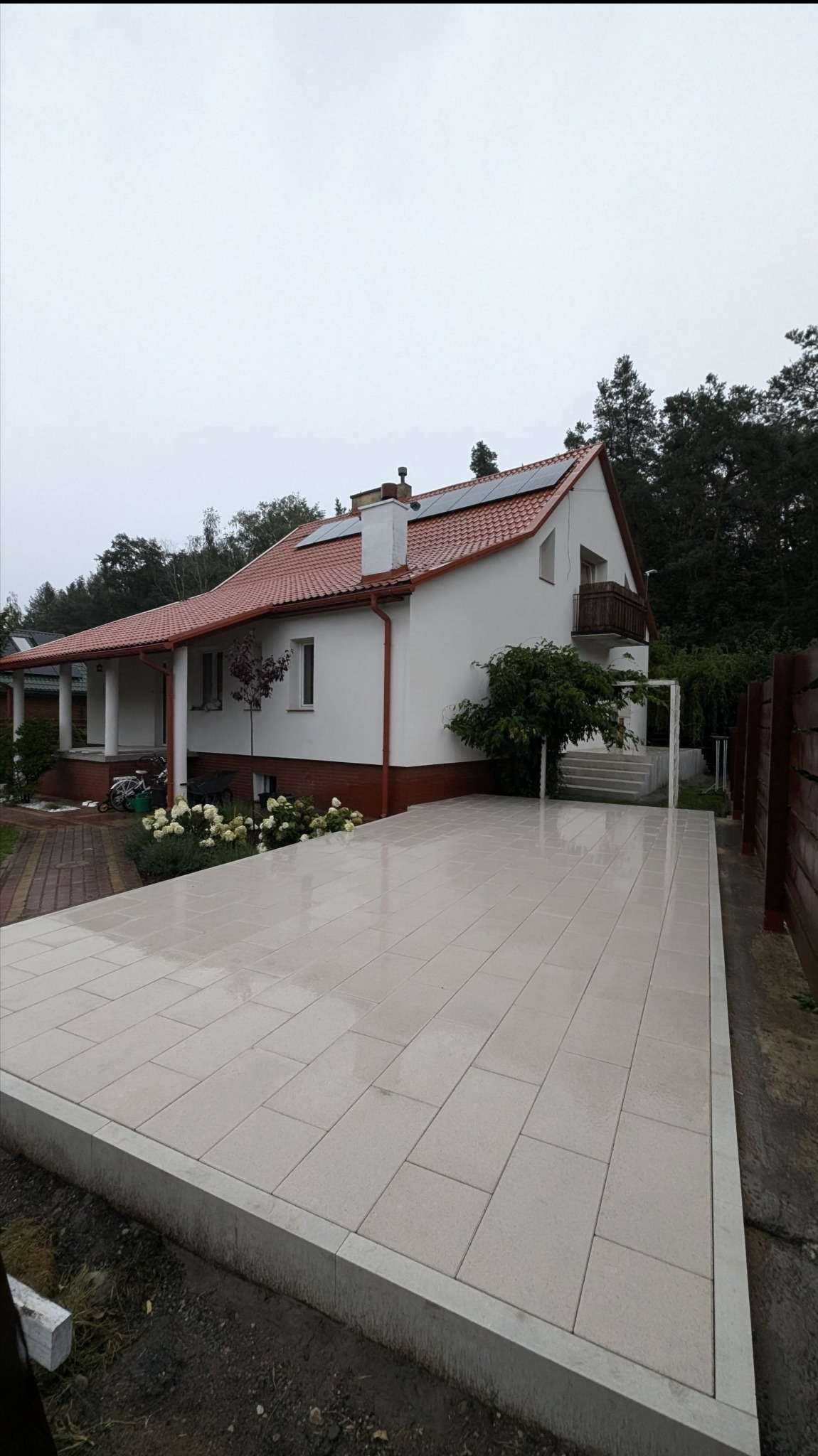 Jasna, nowo ułożona kostka brukowa przed domem z białymi ścianami i czerwoną dachówką, widoczne panele słoneczne na dachu, w tle drzewa.