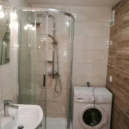 Ukończona łazienka z kabiną prysznicową z zaokrąglonymi drzwiami, umywalką na szafce i pralką w rogu, wykończona płytkami w odcieniach beżu i brązu.