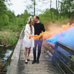 Martyna Branicka-Goździaszek Fotografia - Zakochana para całuje się na drewnianym moście, trzymając w dłoniach dymy w kolorach pomarańczowym i niebieskim, w tle zielony las.