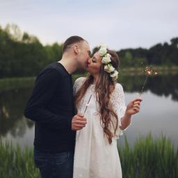 Martyna Branicka-Goździaszek Fotografia - Zakochana para całuje się na tle jeziora w Jarocinie, trzymając w rękach zimne ognie. Dziewczyna ma na głowie wianek z białych kwiatów i koronkową sukienkę.
