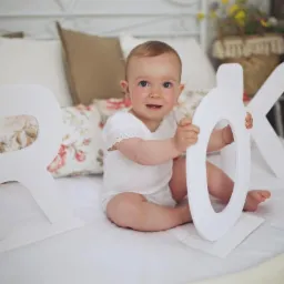 Uśmiechnięte niemowlę siedzi na białej pościeli, trzymając białe litery R, O i K, z dekoracyjnymi poduszkami w tle.