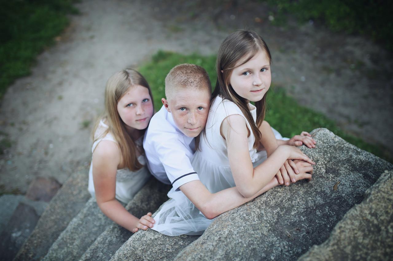 Troje dzieci, dwie dziewczynki i chłopiec, w jasnych ubraniach, siedzą na kamiennych schodach, patrząc w górę w obiektyw.