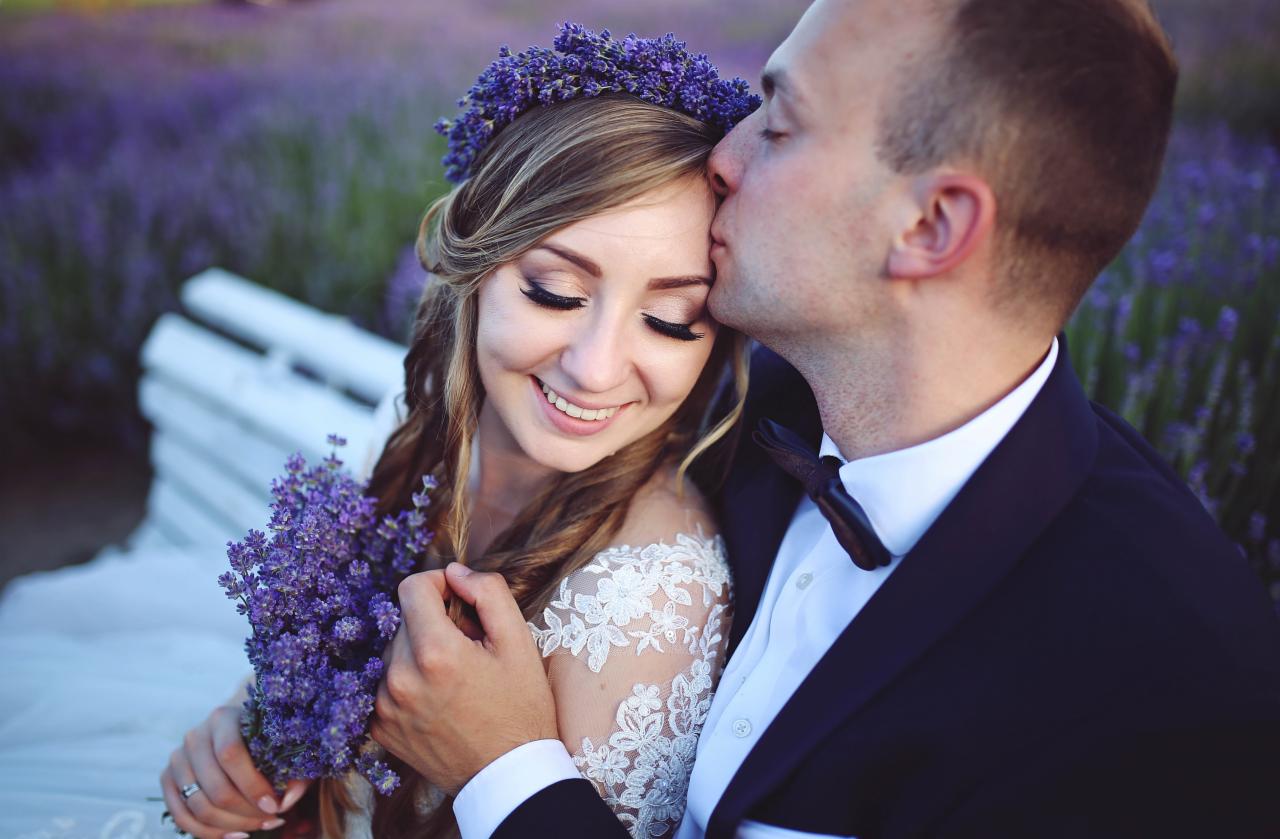 Uśmiechnięta panna młoda w wianku z lawendy, trzyma bukiet lawendy, pan młody całuje ją w czoło na tle kwitnącego pola lawendy.