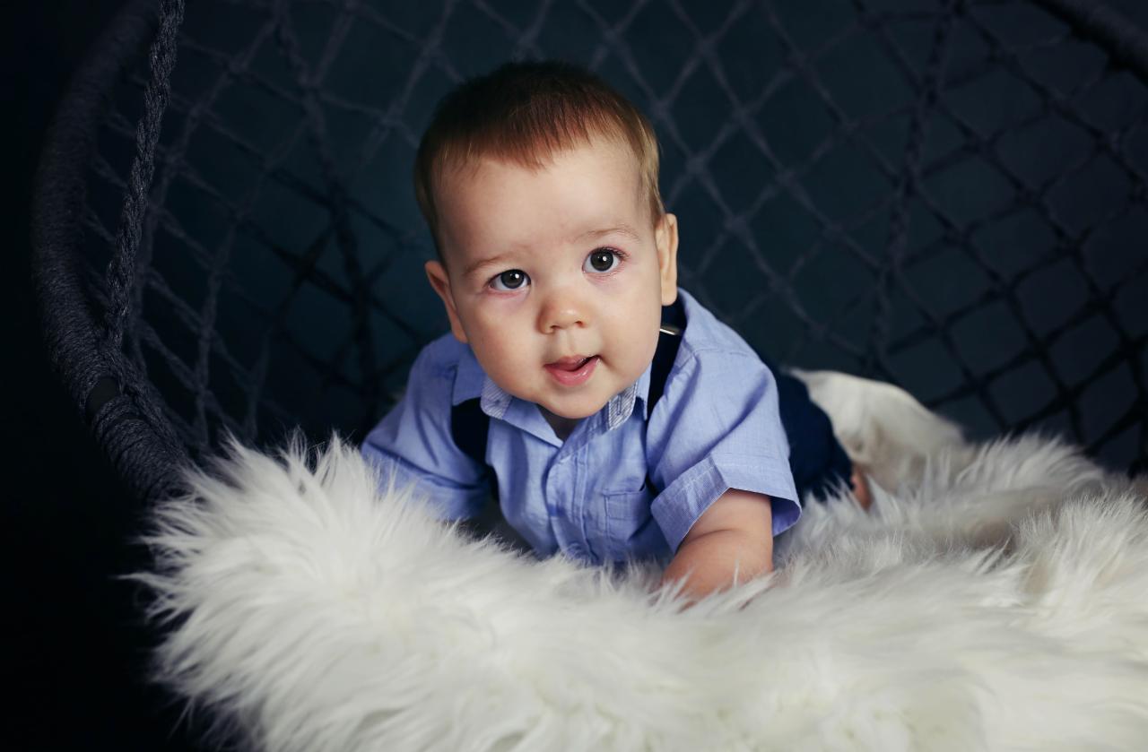 Uroczy niemowlak w błękitnej koszuli i szelkach leży na puszystym białym futrze w wiklinowym koszu na tle ciemnego tła.