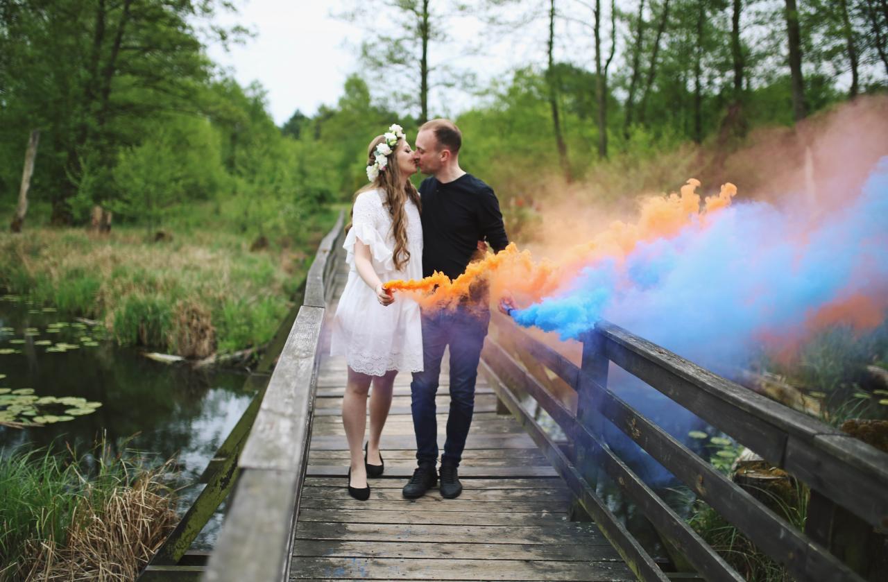 Zakochana para całuje się na drewnianym moście, trzymając w dłoniach dymy w kolorach pomarańczowym i niebieskim, w tle zielony las.