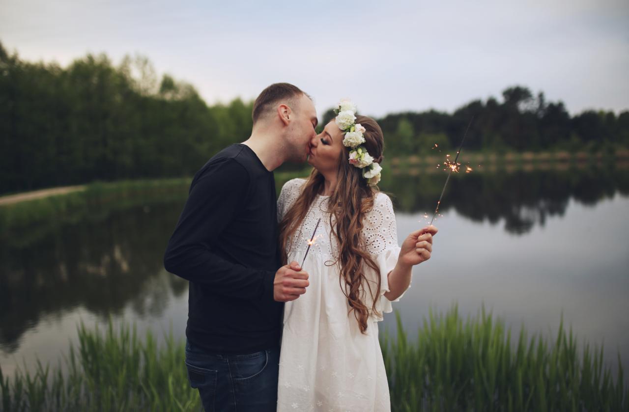 Zakochana para całuje się na tle jeziora w Jarocinie, trzymając w rękach zimne ognie. Dziewczyna ma na głowie wianek z białych kwiatów i koronkową sukienkę.