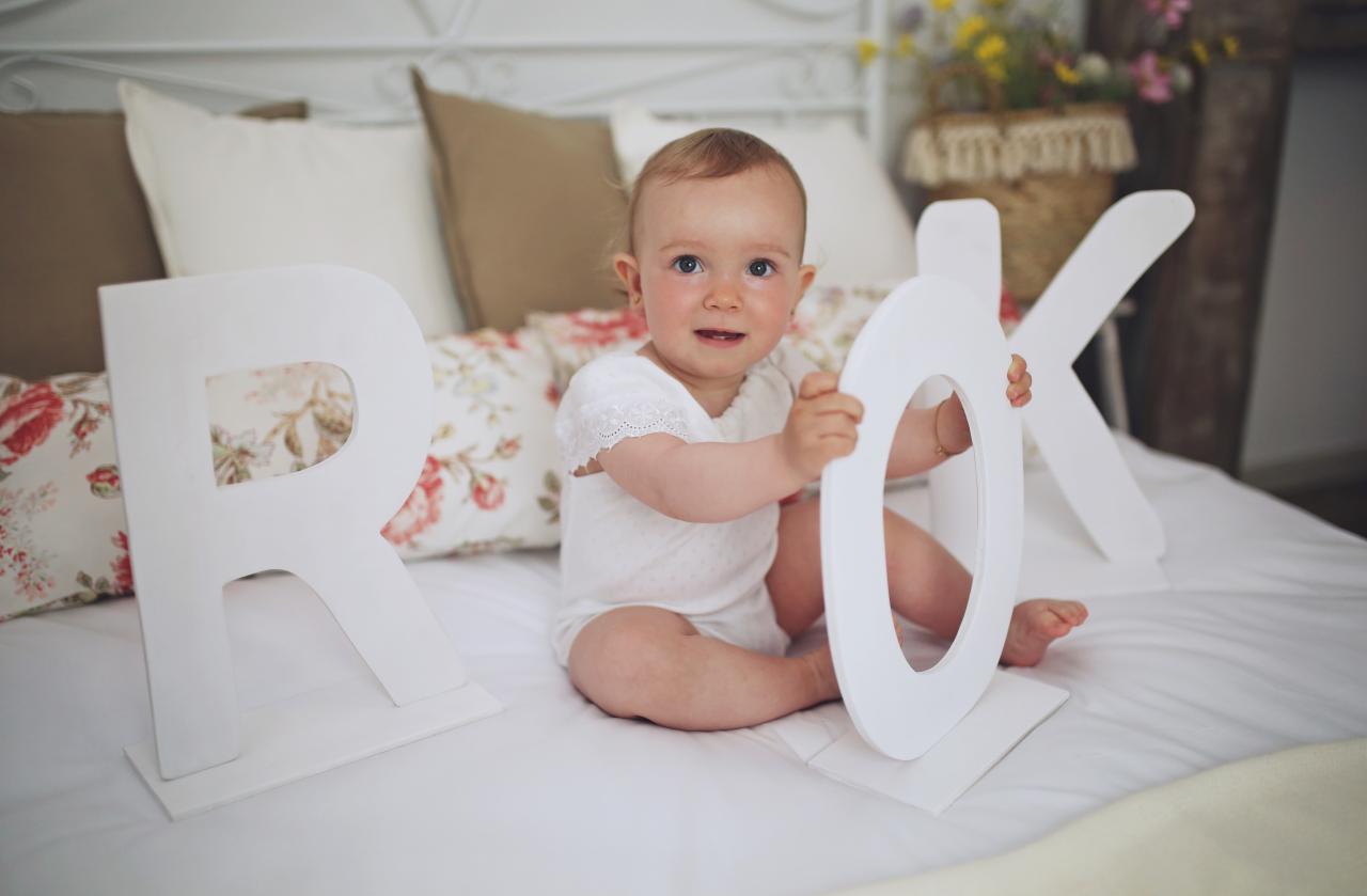 Uśmiechnięte niemowlę siedzi na białej pościeli, trzymając białe litery R, O i K, z dekoracyjnymi poduszkami w tle.