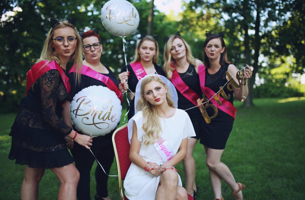 Grupa kobiet w różnym wieku świętuje wieczór panieński w plenerze, z balonami, szarfami i transparentem 'Bride'.