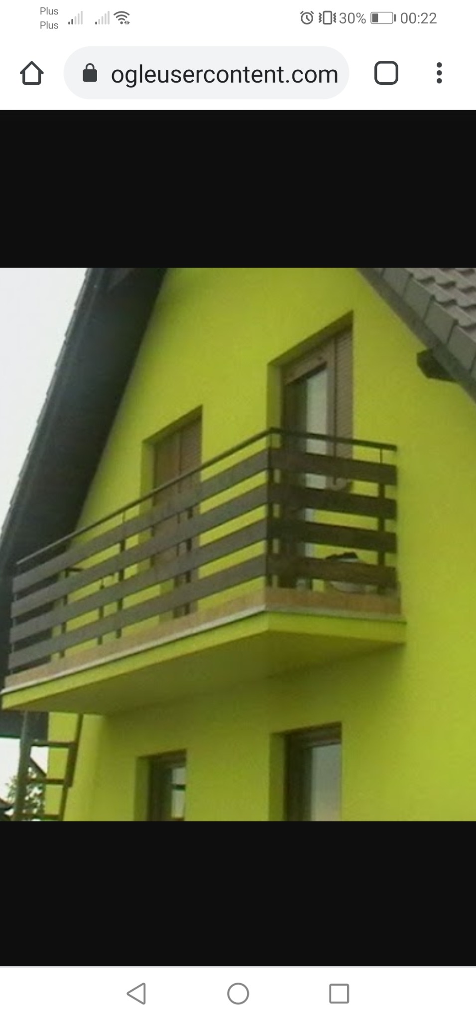 Żółty budynek z drewnianym balkonem i brązową balustradą, widok z dołu, drabina oparta o ścianę z boku balkonu.