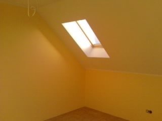 Poddasze po remoncie: żółte ściany, jasne okno dachowe, widoczne gniazdko elektryczne, skośny sufit.