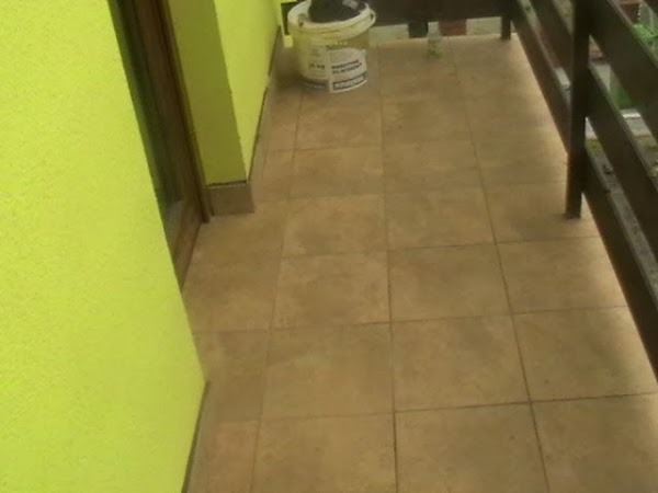 Wyłożony beżowymi płytkami balkon z fragmentem zielonej ściany i brązowej balustrady; wiadro z klejem w rogu.