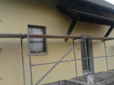 Elewacja domu w trakcie malowania na żółto, widoczne rusztowanie, okno z firanką i fragment drewnianej konstrukcji dachu