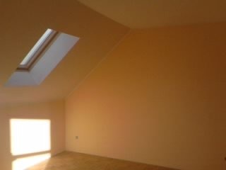 Pomalowane na beżowo poddasze ze skośnym sufitem i oknem dachowym, widok na fragment podłogi i gładkie ściany.