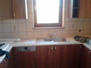 Kuchnia w trakcie remontu: widoczne szafki z ciemnego drewna, biały blat, zlewozmywak z baterią, okno z drewnianą ramą oraz białe kafelki na ścianie. Część blatu przykryta folią ochronną.