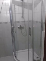 Kabina prysznicowa narożna z chromowanymi profilami i uchwytami, widoczna na tle białych płytek ceramicznych z delikatnym wzorem.