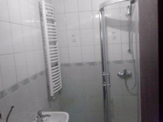 Biała łazienka z kabiną prysznicową z zaokrąglonymi drzwiami, grzejnikiem drabinkowym i umywalką. Widoczne białe płytki na ścianach z delikatnym wzorem.