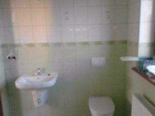 Umywalka i toaleta w łazience z białymi płytkami i zielonym pasem dekoracyjnym, widoczne niedoskonałości w wykończeniu ścian.