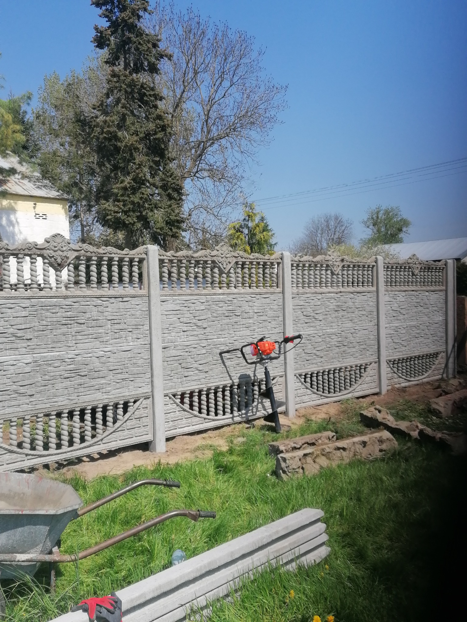 Montaż prefabrykowanego betonowego ogrodzenia z ozdobnymi elementami, widoczny wiertniczy silnik spalinowy, taczka i stos słupków ogrodzeniowych na trawie.