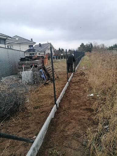 Montaż ogrodzenia panelowego na betonowej podmurówce, widoczna osoba przy pracy, w tle budynki mieszkalne i teren zarośnięty trawą.