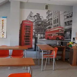 Sala lekcyjna z fototapetą przedstawiającą londyńską ulicę z czerwonym autobusem i budką telefoniczną, tablicą z alfabetem i rzędami pomarańczowych krzeseł przy drewnianych stolikach.