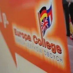 Pomarańczowa tablica reklamowa Europa College z flagami: niemiecką, brytyjską i hiszpańską, wskazująca kierunek szkoły językowej.