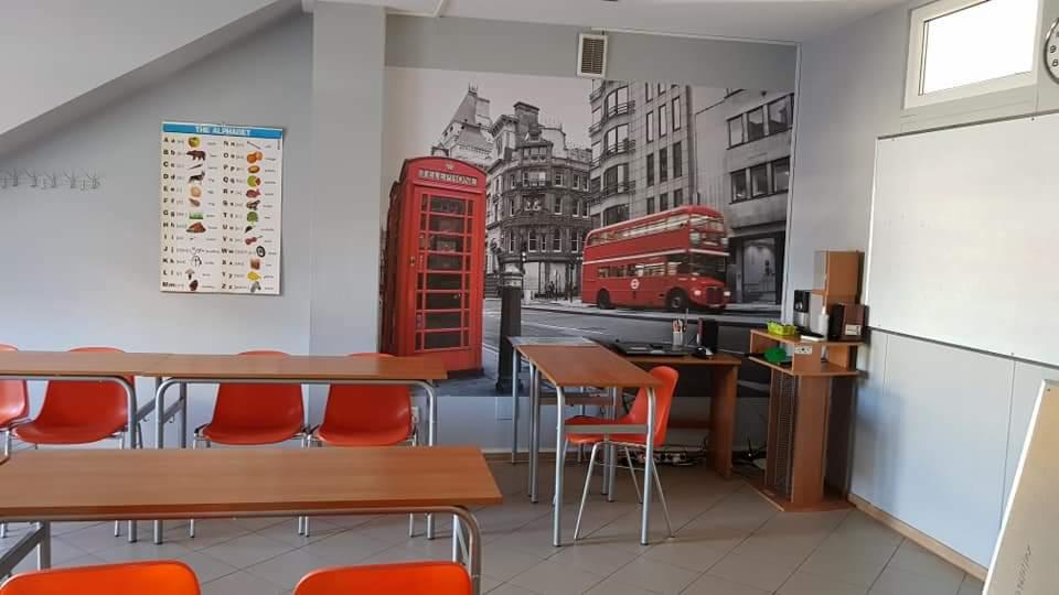 Sala lekcyjna z fototapetą przedstawiającą londyńską ulicę z czerwonym autobusem i budką telefoniczną, tablicą z alfabetem i rzędami pomarańczowych krzeseł przy drewnianych stolikach.