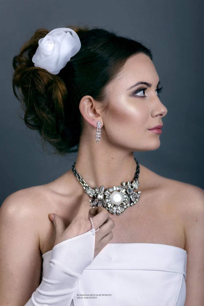 Portret kobiety z upiętymi włosami ozdobionymi białą różą, w bogato zdobionym naszyjniku i kolczykach, z delikatnym makijażem oczu.