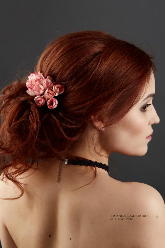 Profil kobiety z rudymi włosami upiętymi w kok, ozdobionymi spinką z różowymi kwiatami, delikatny makijaż oczu, czarny choker na szyi, widoczne plecy.