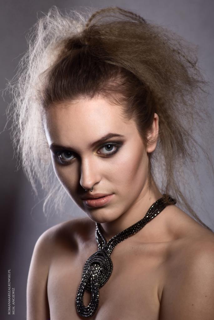 Portret kobiety z ciemnym makijażem oczu, piercingiem w nosie i ekstrawagancką fryzurą typu up-do, ubrana w ozdobny naszyjnik.