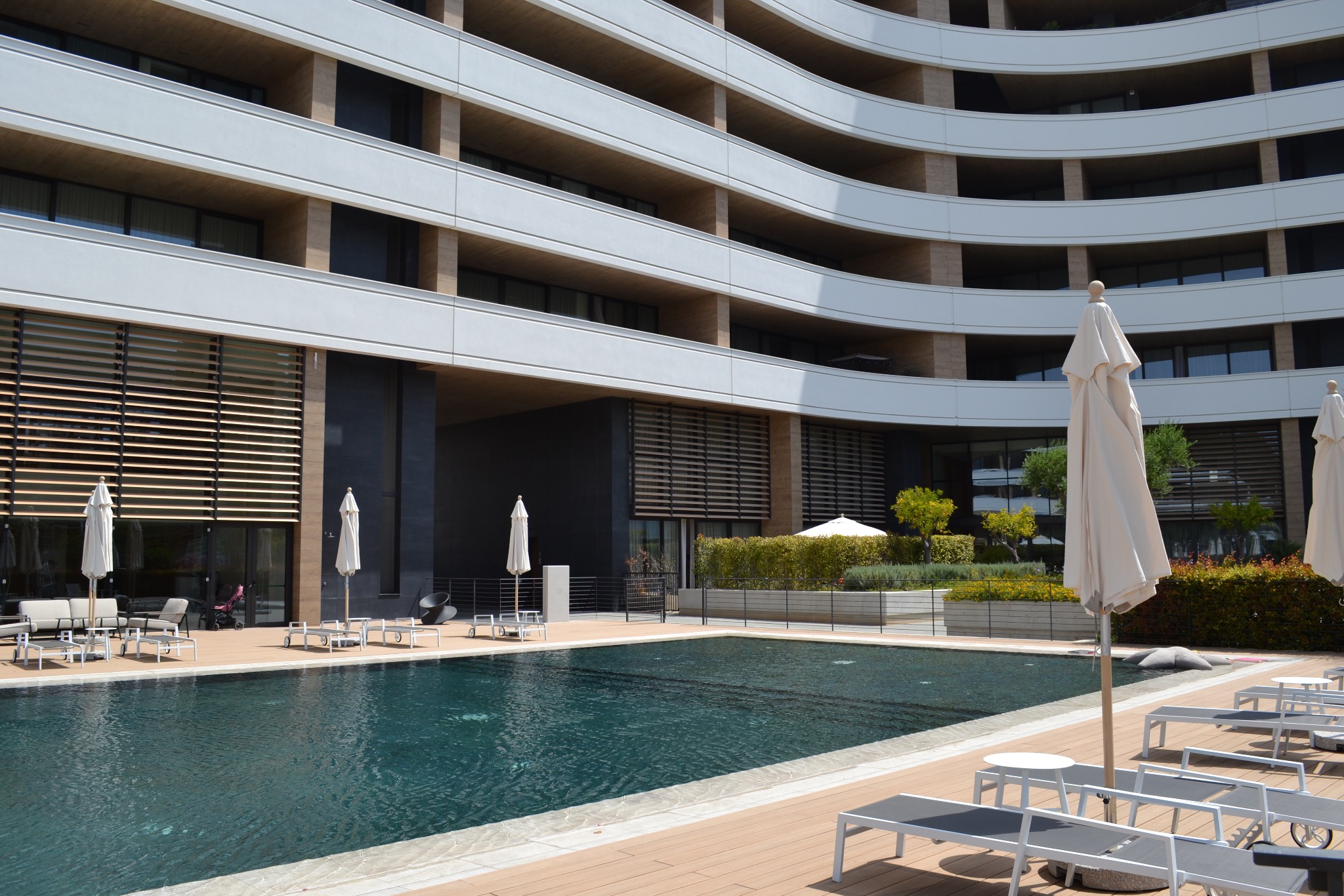 Luksusowy apartamentowiec z basenem i leżakami na tarasie. Nowoczesna architektura z zaokrąglonymi balkonami i drewnianymi żaluzjami.