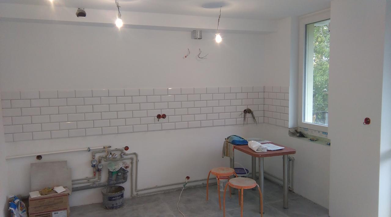 Wnętrze w trakcie prac wykończeniowych: białe kafelki ułożone do połowy ściany, widoczne instalacje hydrauliczne, okno z widokiem na zieleń, dwa taborety i stół z narzędziami.